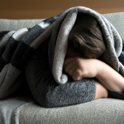 description: a person lying on a couch with a blanket over them, holding their head in their hands and looking miserable.